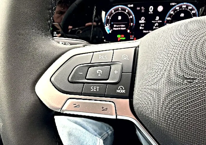 Gros plan sur le volant noir de la Volkswagen Golf 2026 avec commandes de régulateur adaptatif et tableau de bord digital en arrière-plan.