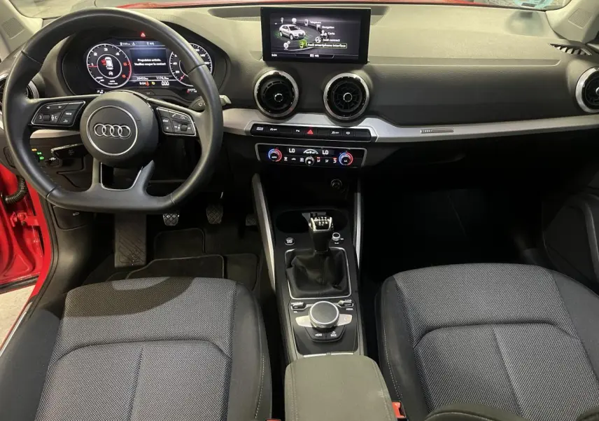 Intérieur Audi Q2 2024 rouge, vue de face sur tableau de bord, volant sport cuir, écran central et boîte manuelle.