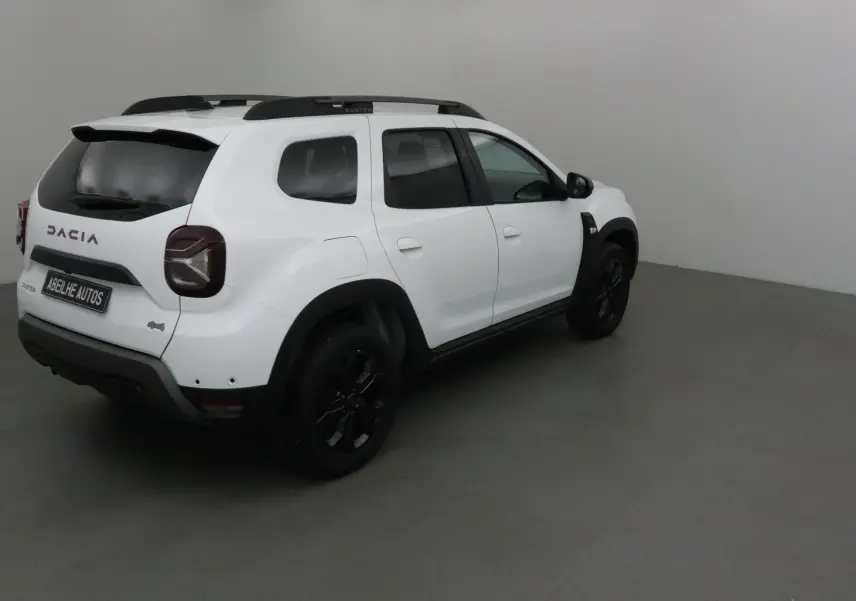 Dacia Duster 4x4 blanc en 3/4 arrière droit, avec jantes noires et barres de toit visibles dans un intérieur neutre.