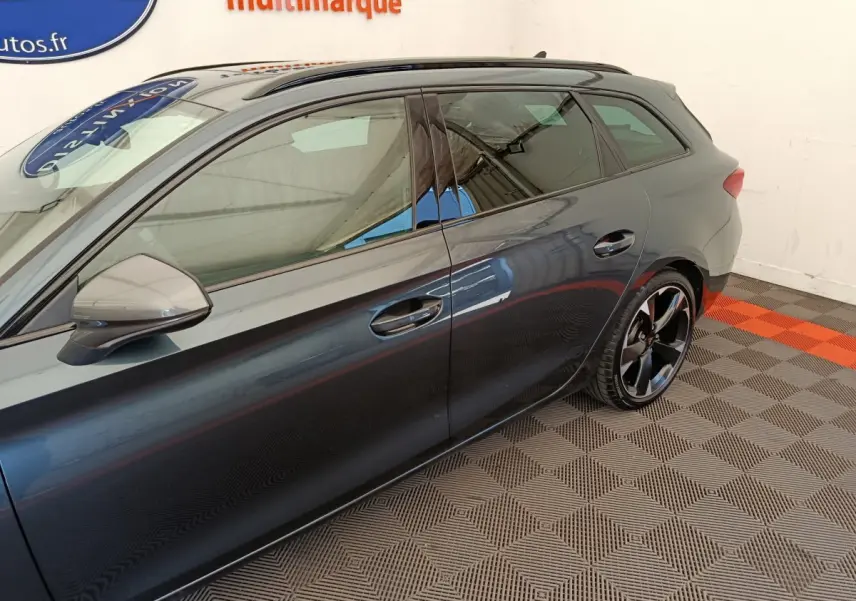 Vue latérale côté gauche d'une CUPRA LEON ST gris foncé avec jantes noires et toit ouvrant visible en intérieur showroom.