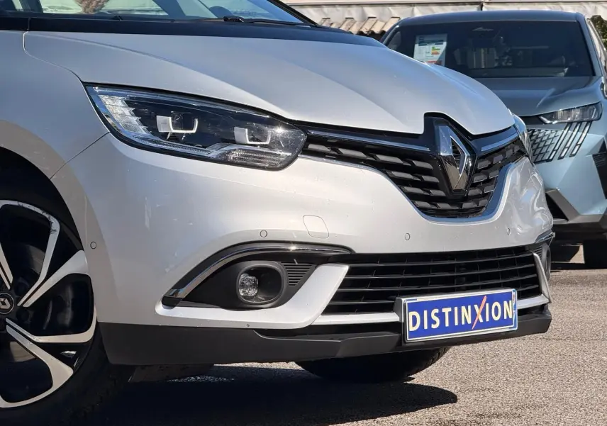Vue 3/4 avant d'un Renault Scénic IV gris platine avec jantes noires et logo Distinxion sur le pare-chocs.