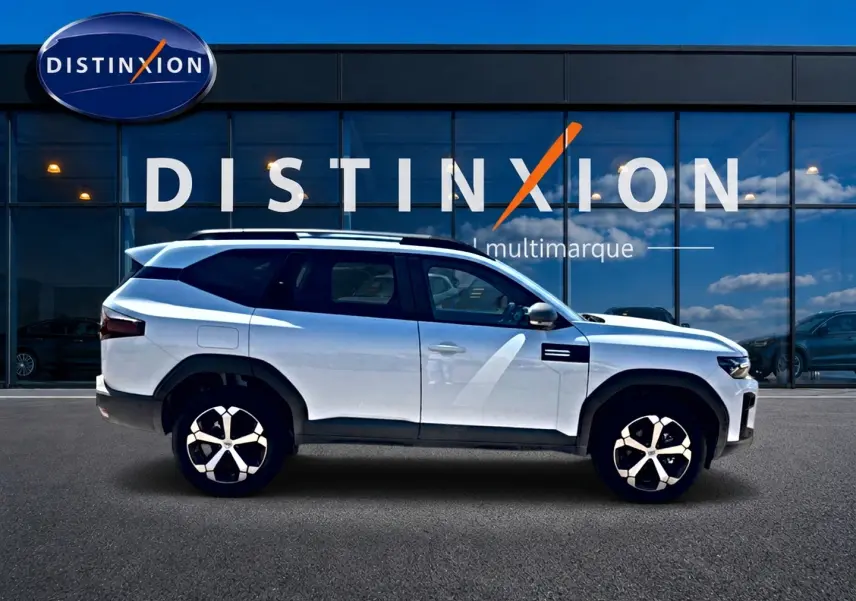 Profil droit du Dacia Bigster blanc 2026 avec jantes alu et barres de toit devant une vitrine Distinxion multimarque.
