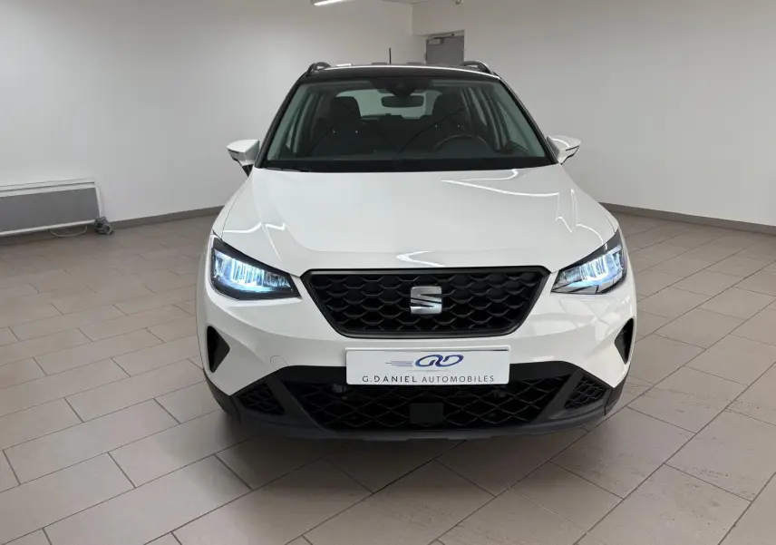 Vue de face d'une SEAT Arona 2023 blanche avec toit noir, phares allumés dans un intérieur neutre.