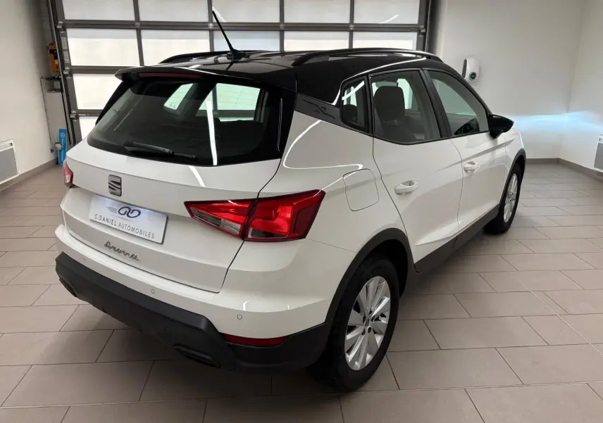 Vue 3/4 arrière droite du SEAT Arona blanc avec toit noir, soulignant son design compact et élégant en intérieur showroom.