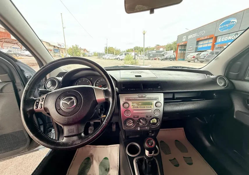 Intérieur noir de la Mazda2 2004, vue du poste de conduite avec volant et levier de vitesses manuelle visibles.