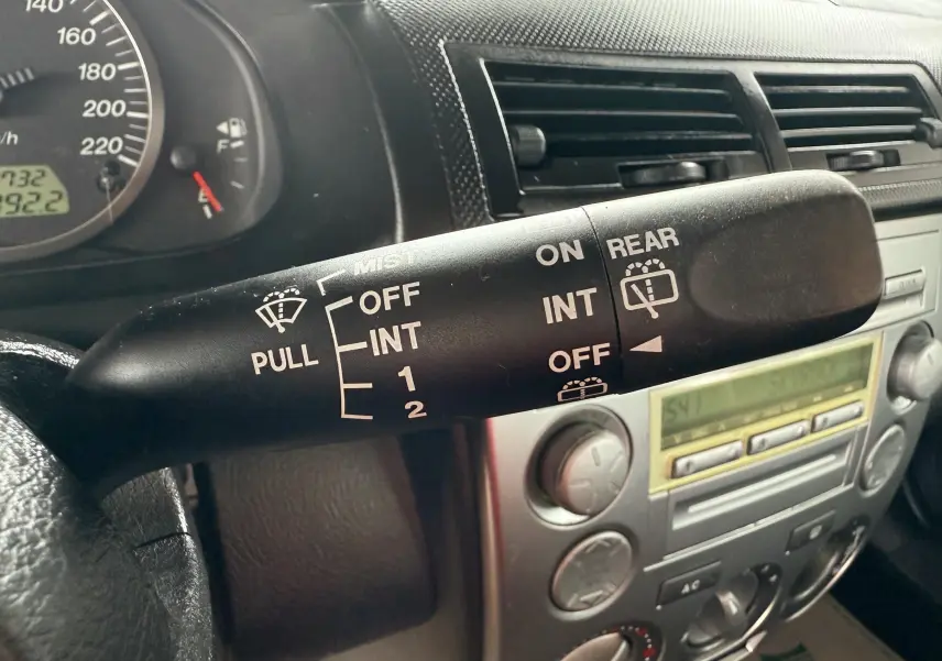 Gros plan sur la manette d'essuie-glace côté gauche du tableau de bord d'une Mazda2 gris 2004, avec commandes visibles.