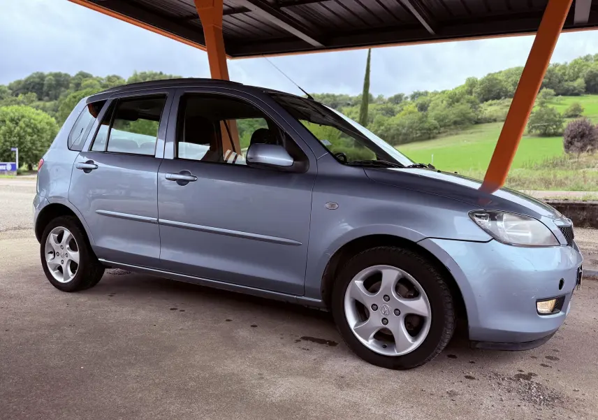 Vue de profil côté gauche d'une Mazda2 gris clair 5 portes garée sous un abri avec un paysage verdoyant en arrière-plan.