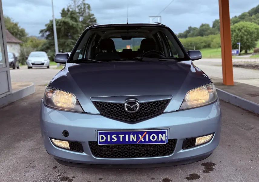 Vue frontale d'une Mazda2 gris clair avec phares allumés et plaque d'immatriculation Distinxion visible.