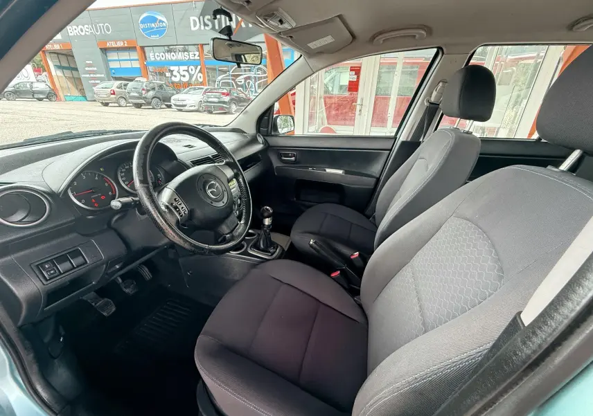Intérieur avant droit de la Mazda2 1.4 TDCI 2004 avec sièges tissu noir et volant multifonction.