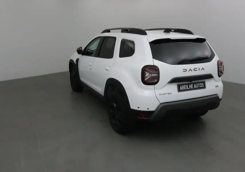 Vue 3/4 arrière gauche d'un Dacia Duster blanc 2024 avec barres de toit et jantes noires dans un studio.