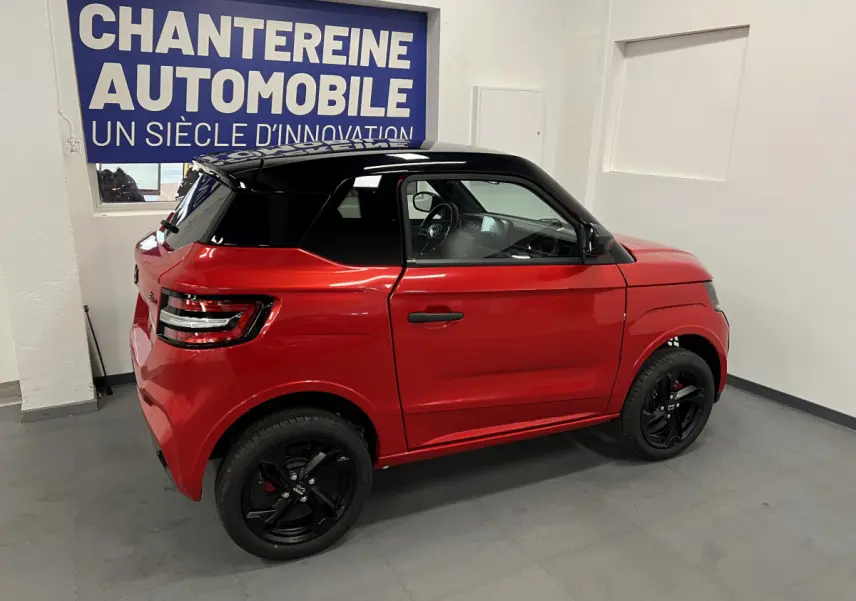 Vue côté droit arrière de la LIGIER JS50 BEV 8KWH ELITE rouge avec toit noir et jantes alu noires en intérieur showroom.