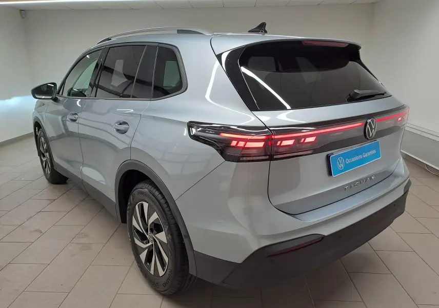 Vue 3/4 arrière droite du Volkswagen Tiguan 2025 en argent Dolomite avec feux arrière LED allumés dans un intérieur showroom.