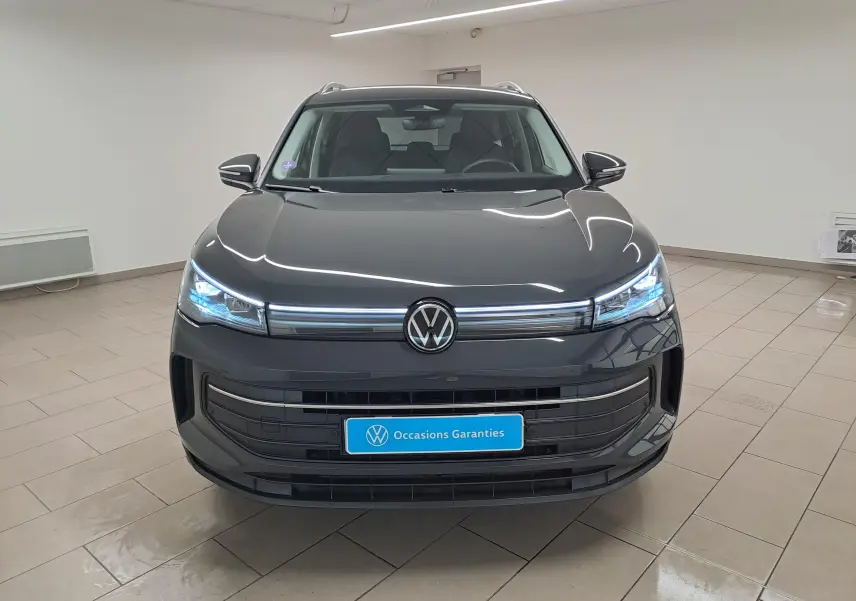 Vue frontale d'un Volkswagen Tiguan 2024 gris Urano avec phares allumés et plaque "Occasions Garantis" en intérieur.