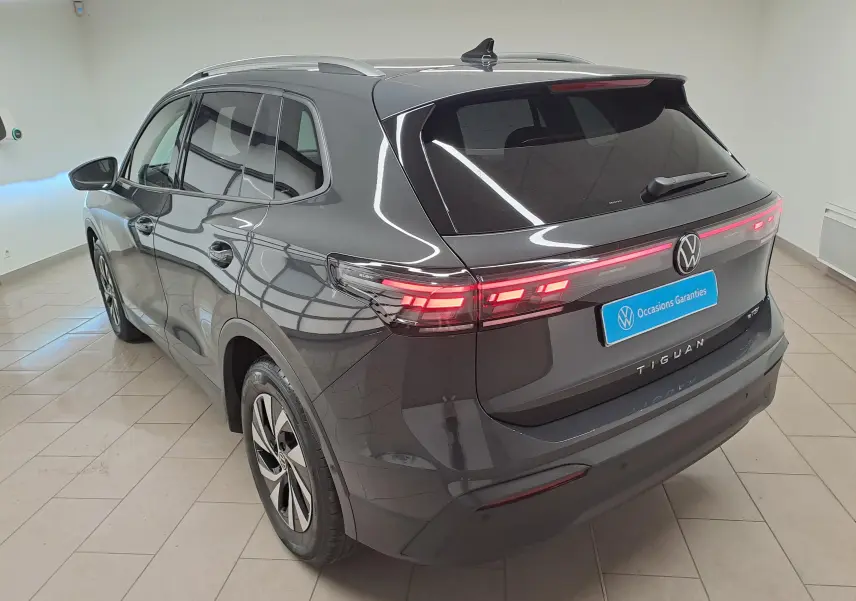 Vue 3/4 arrière droite du Volkswagen Tiguan 2024 gris Urano avec feux arrière allumés et crochet d'attelage escamotable visible.