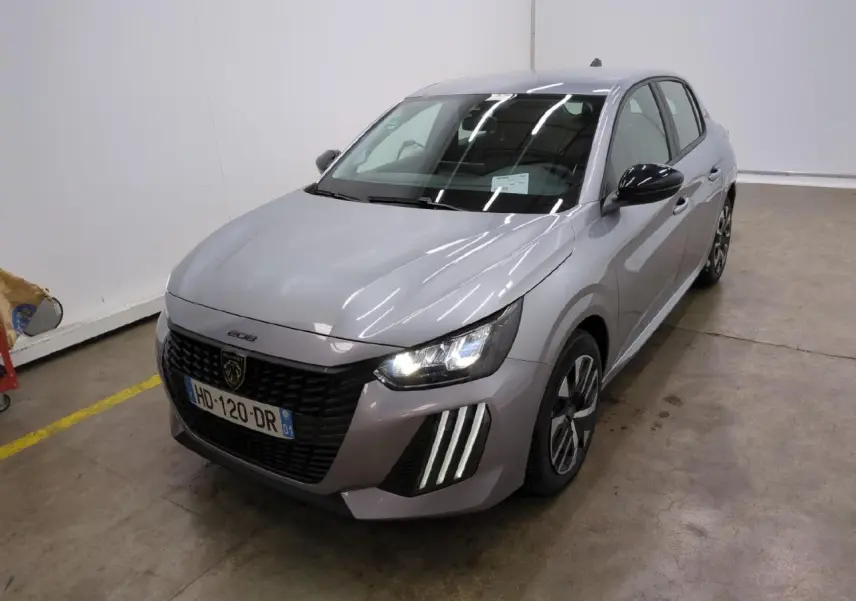 Peugeot 208 gris clair vue 3/4 avant droit avec calandre noire et feux LED allumés dans un garage.