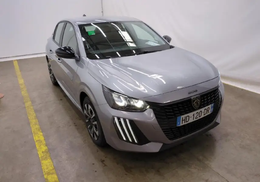 Peugeot 208 gris clair vue 3/4 avant droit avec calandre noire et feux LED allumés dans un garage.