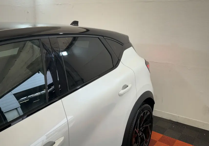 Vue côté gauche arrière du Renault Captur blanc avec toit noir et jantes noires sportives dans un intérieur lumineux.