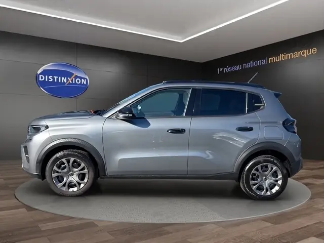Vue de profil côté gauche d'une Citroën C3 gris foncé 2025 avec spoiler de coffre, sur fond intérieur showroom.