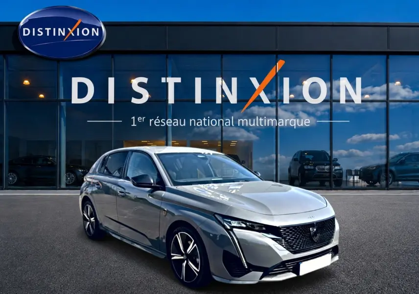 Peugeot 308 gris en 3/4 avant droit, avec calandre noire et jantes alliage distinctives, devant un showroom Distinxion.