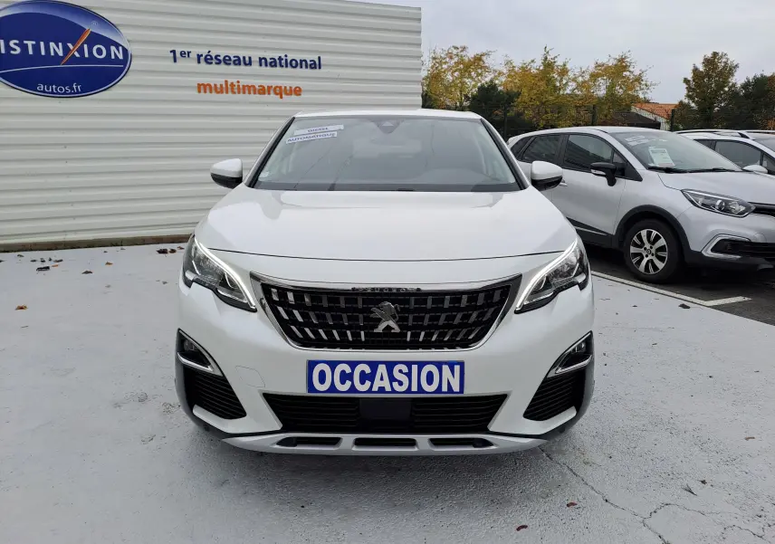 Vue avant d'un Peugeot 3008 blanc avec plaque "OCCASION" sur un parking extérieur devant un panneau publicitaire.