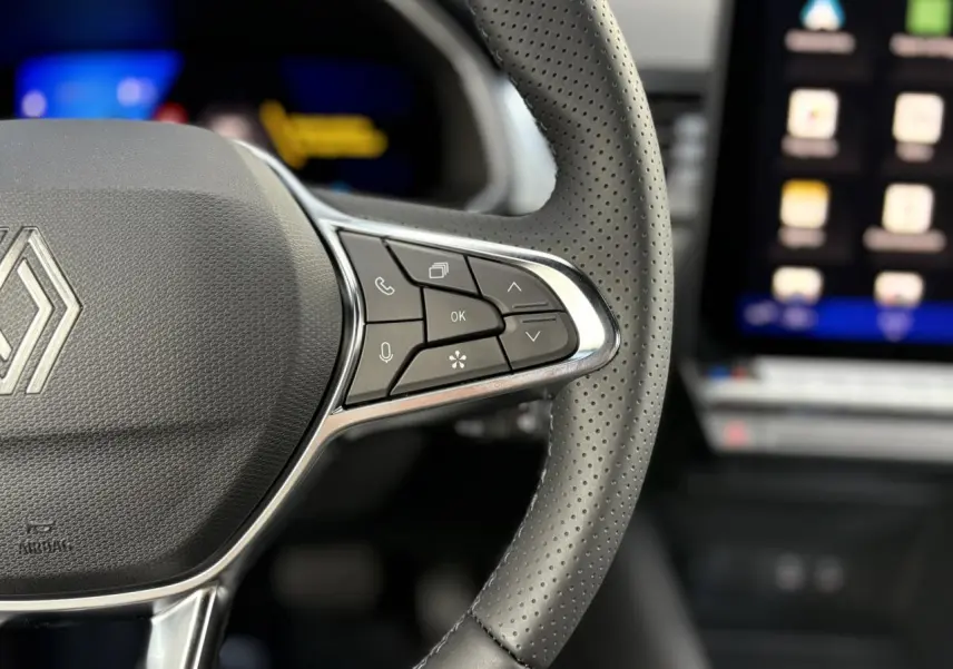 Gros plan sur le volant en cuir perforé de la Renault SYMBIOZ noir étoile, avec commandes multifonctions et écran tactile flou en arrière-plan.