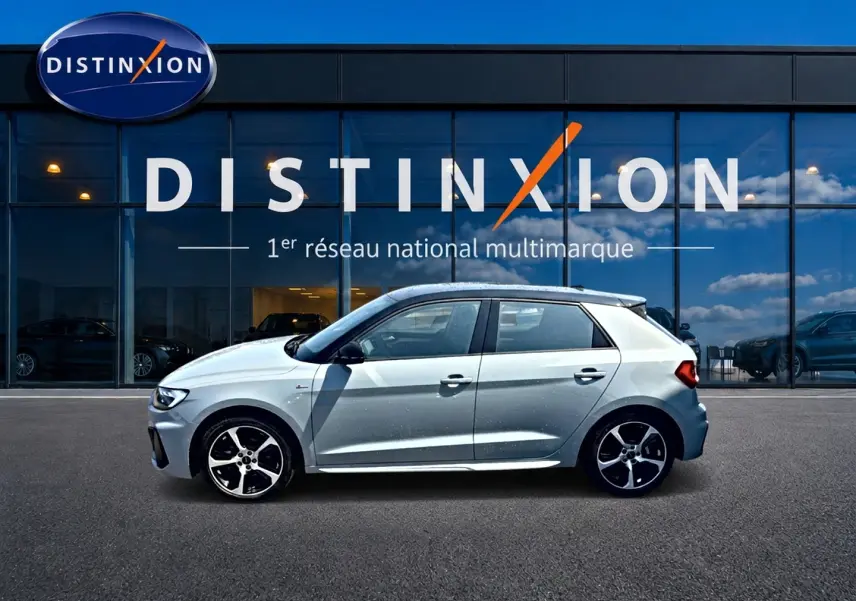 Profil droit d'une Audi A1 Sportback 30 TFSI S line gris arrow avec jantes alliage noires devant une vitrine Distinxion.