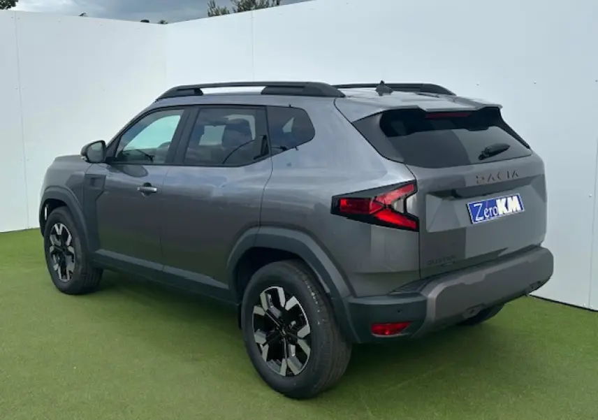 Vue 3/4 arrière droite du Dacia Duster III gris schiste métallisé avec barres de toit noires et jantes alliage 17 pouces.