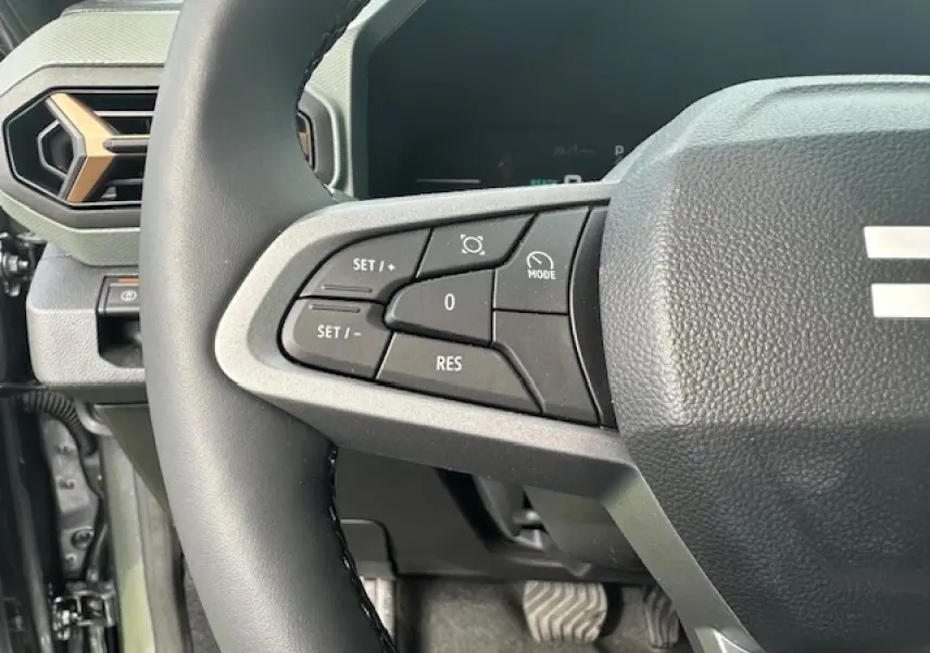 Gros plan sur les commandes au volant côté gauche du Dacia Duster III gris schiste, avec tableau de bord digital en arrière-plan.