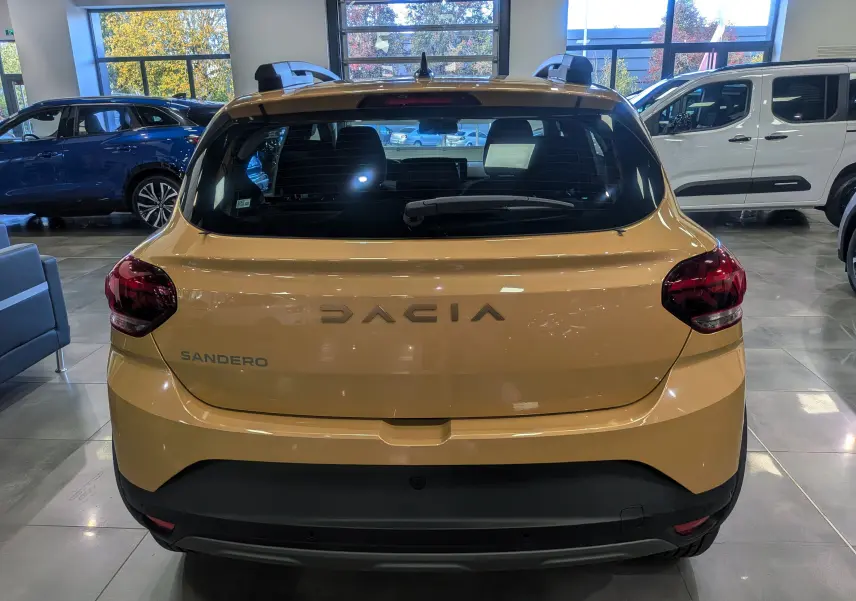 Vue arrière d'une Dacia Sandero Stepway beige Safari 2025 en showroom, avec feux arrière foncés et toit avec barres de toit.