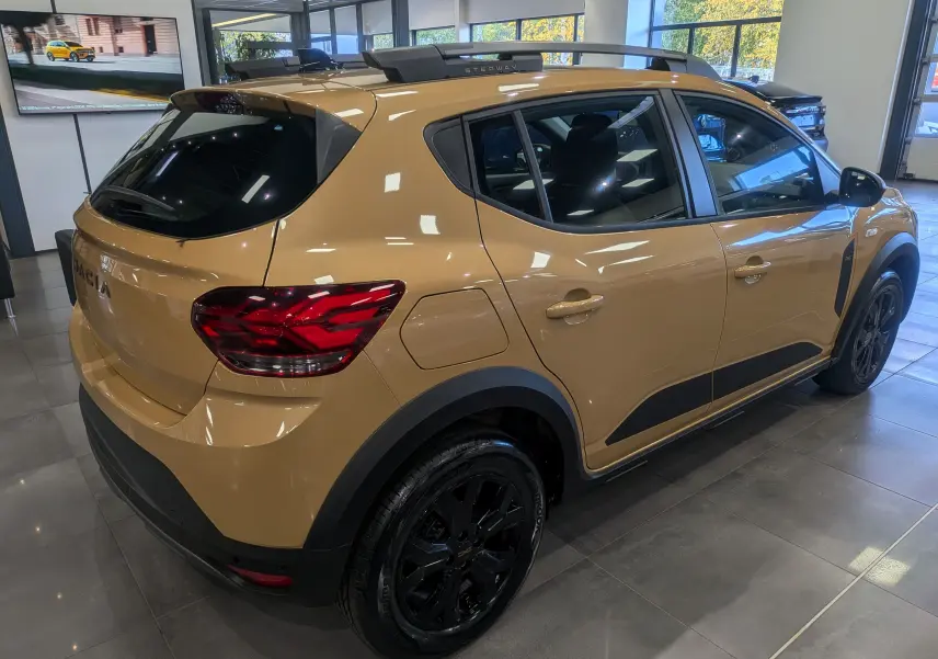 Vue 3/4 arrière droite d'une Dacia Sandero Stepway beige Safari avec jantes noires et barres de toit Stepway visibles.