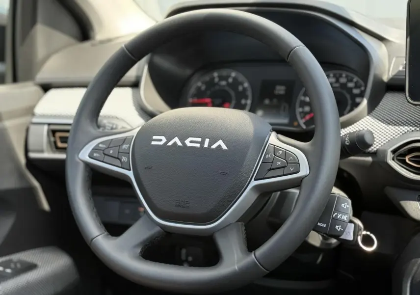Gros plan sur le volant noir Dacia avec commandes intégrées, tableau de bord et partie de la console centrale visibles.