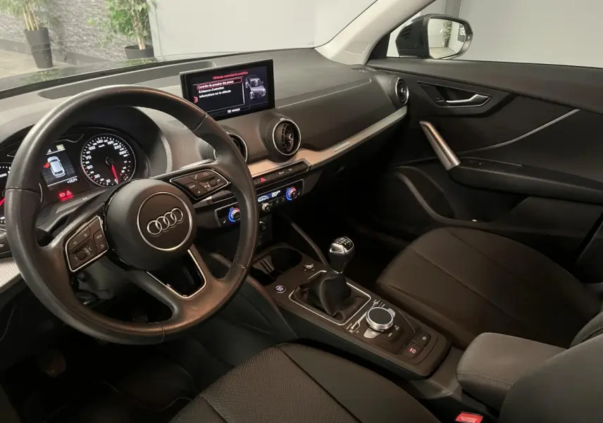 Intérieur Audi Q2 2023 vu côté conducteur, volant cuir, tableau de bord noir et levier de vitesse manuel.