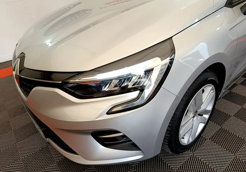 Vue 3/4 avant droit d'une Renault Clio Business TCe 100 gris clair, mettant en valeur ses phares LED et la calandre noire brillante.