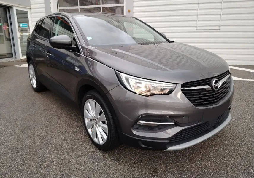 Opel Grandland X gris platinium en 3/4 avant droit, avec phares allumés et jantes alliage visibles.