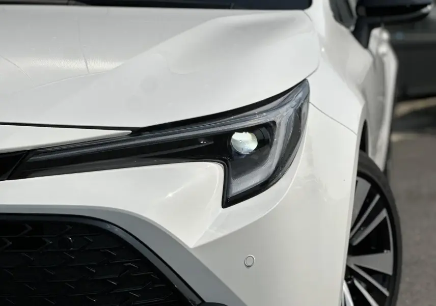Gros plan sur le phare avant droit d'une Toyota Corolla Touring Sport blanche Neige, avec jante alliage visible.