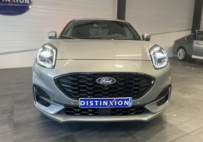 Vue frontale d'un Ford Puma gris clair avec calandre noire hexagonale et phares LED allumés en intérieur showroom.