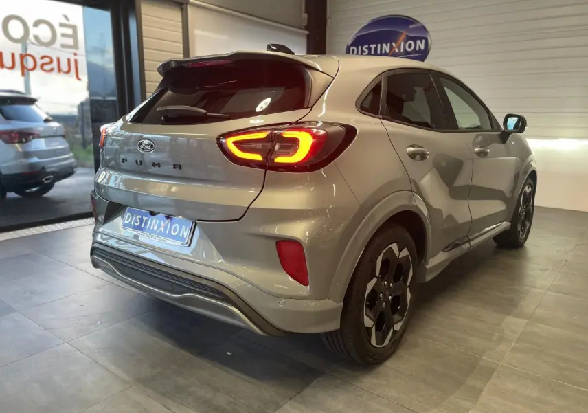 Vue 3/4 arrière droite d'un Ford Puma gris clair avec feux arrière allumés en intérieur showroom.