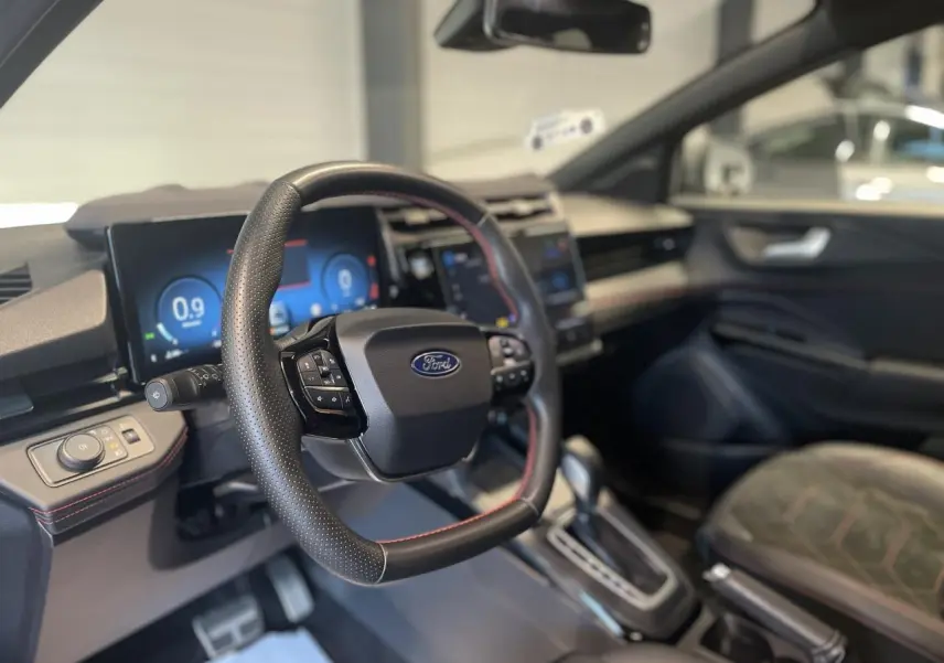 Vue intérieure côté conducteur du Ford Puma 2024, volant cuir noir avec surpiqûres rouges et tableau de bord numérique.