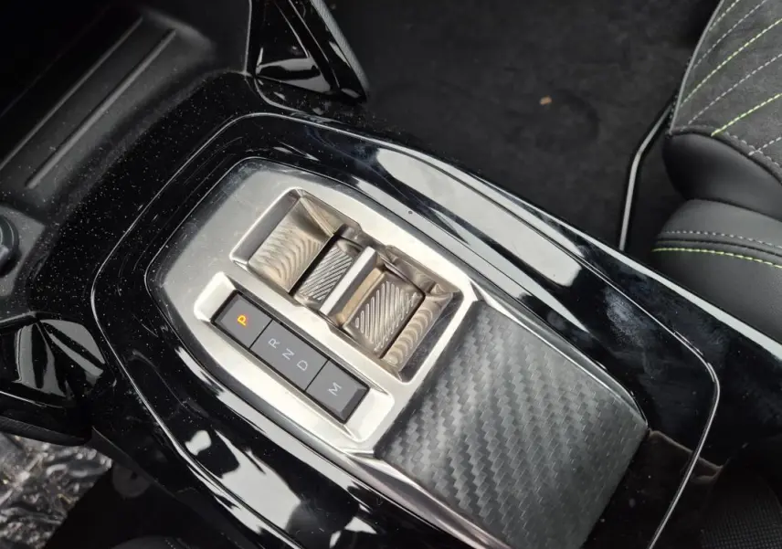 Gros plan sur la console centrale noire brillante de la Peugeot 208 Hybrid 110 e-DCS6 GT avec levier de vitesses électrique et finition carbone.