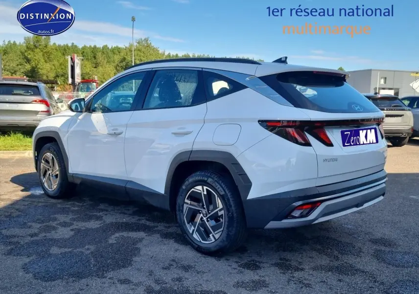 Vue 3/4 arrière droite du Hyundai Tucson blanc atlas avec jantes alliage et barres de toit noires sur parking extérieur.