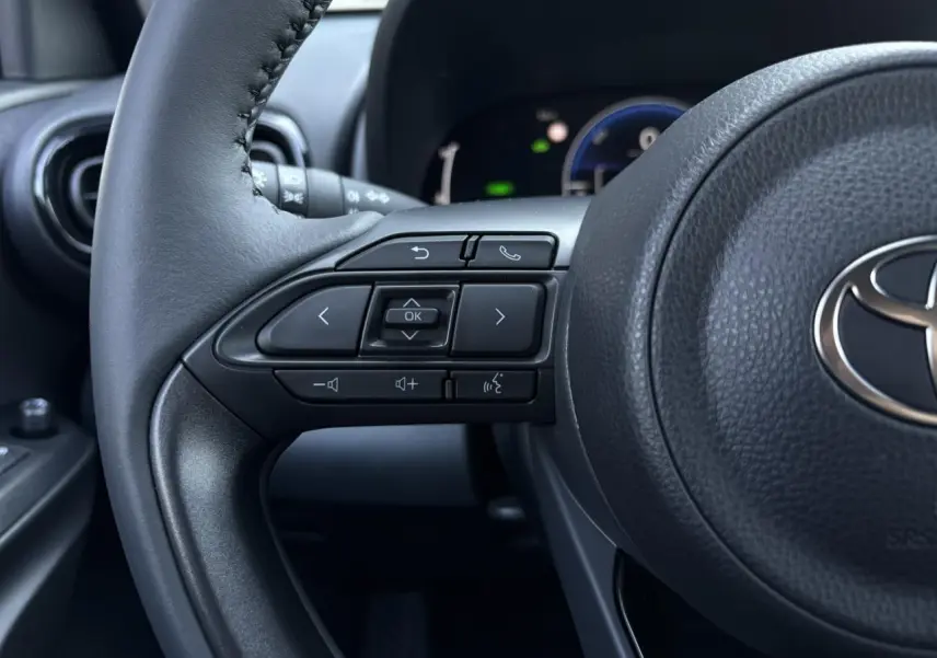Gros plan sur le côté gauche du volant cuir noir de la Toyota Yaris 2025, avec commandes intégrées et tableau de bord numérique en arrière-plan.