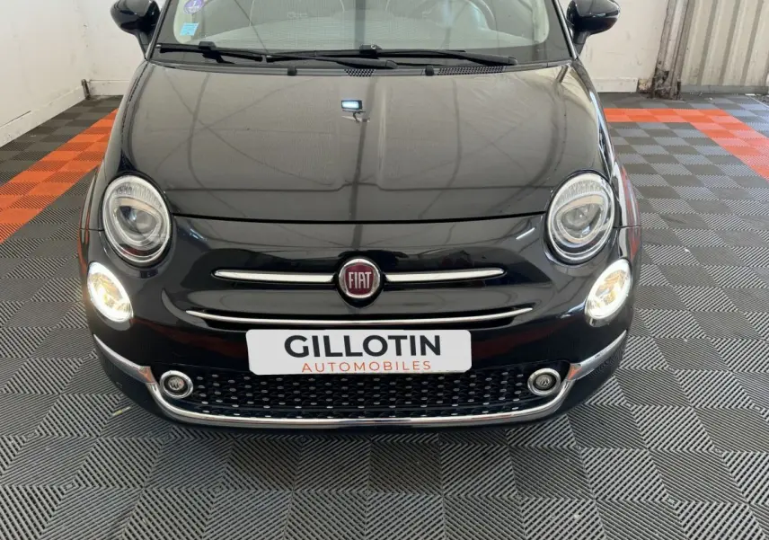 Vue frontale d'une Fiat 500 noire avec phares allumés et calandre chromée dans un garage.