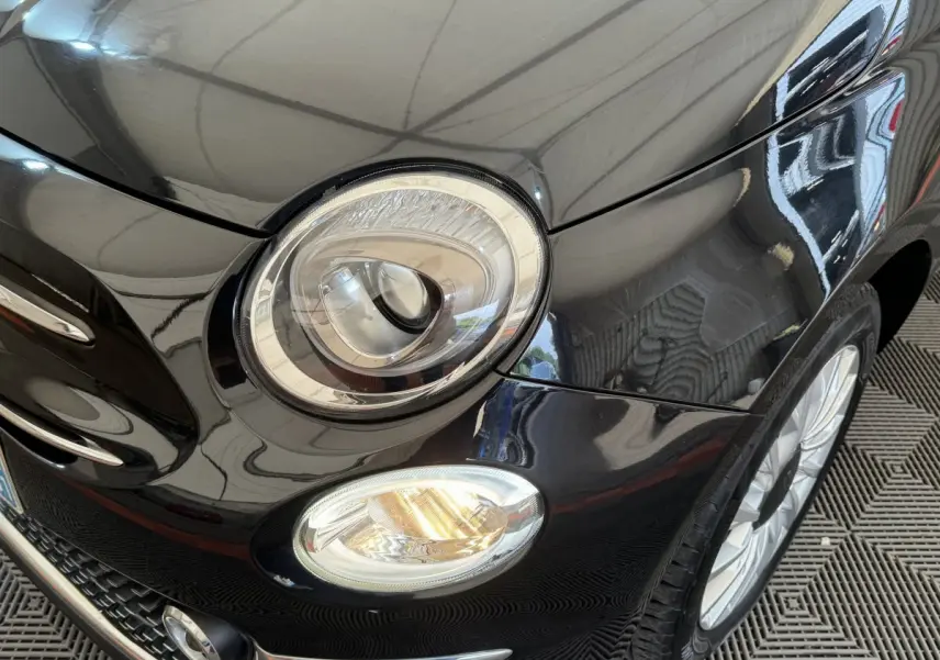 Gros plan sur l'avant droit noir brillant de la Fiat 500 1.2 8V 69ch Lounge avec phare rond et antibrouillard allumé.