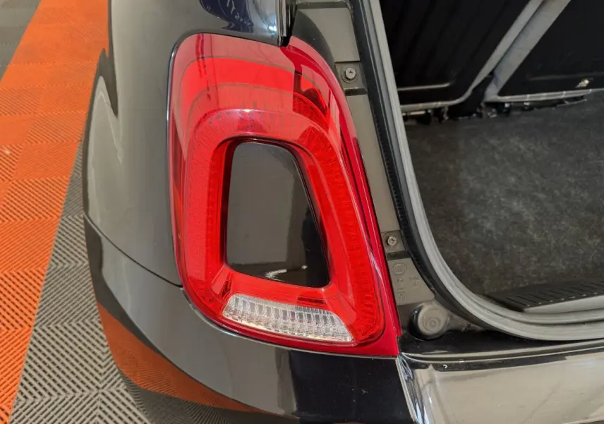 Gros plan sur le feu arrière droit rouge et blanc d'une FIAT 500 noire avec coffre ouvert sur sol carrelé orange et gris.