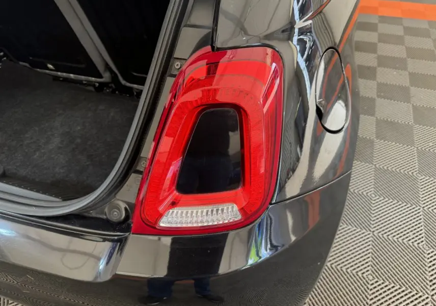 Gros plan sur le feu arrière droit rouge d'une Fiat 500 noire avec coffre ouvert, sur sol à damier gris et orange.
