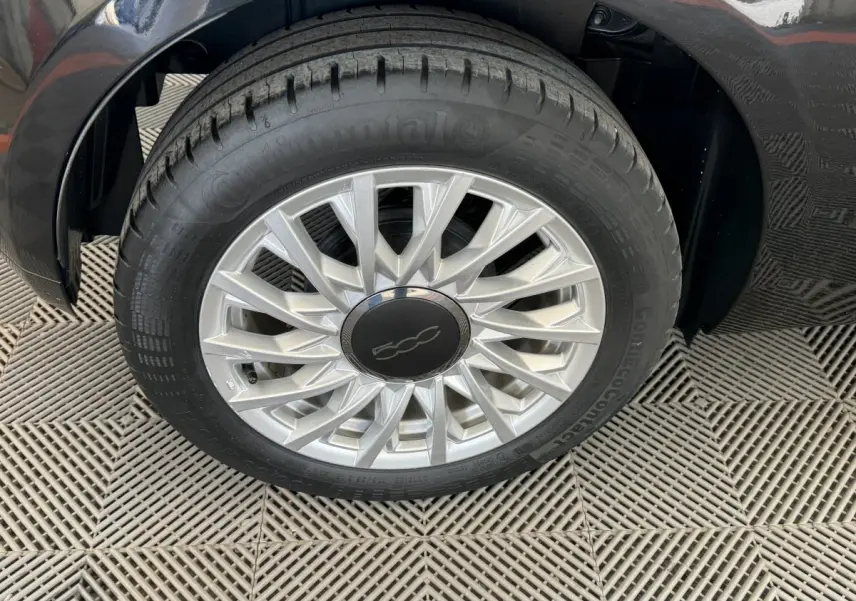 Gros plan sur la roue avant gauche noire d'une Fiat 500 1.2 8V 69ch Lounge avec jante argentée à rayons fins.