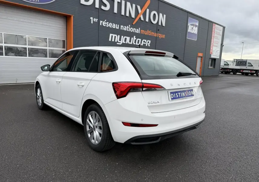 Vue 3/4 arrière droite d'une Skoda Scala blanche 2023 avec jantes alliage et feux arrière distinctifs, stationnée devant un bâtiment.