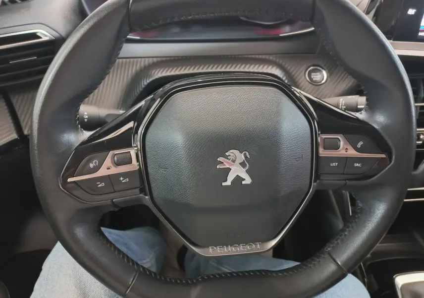 Vue rapprochée du volant cuir noir de la Peugeot 208 PureTech 100 S&S Allure avec commandes intégrées et logo lion central.