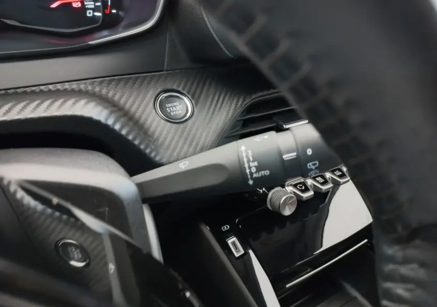 Gros plan sur le tableau de bord carbone et le bouton start/stop d'une Peugeot 208 blanche, version PureTech 100 S&S Allure.
