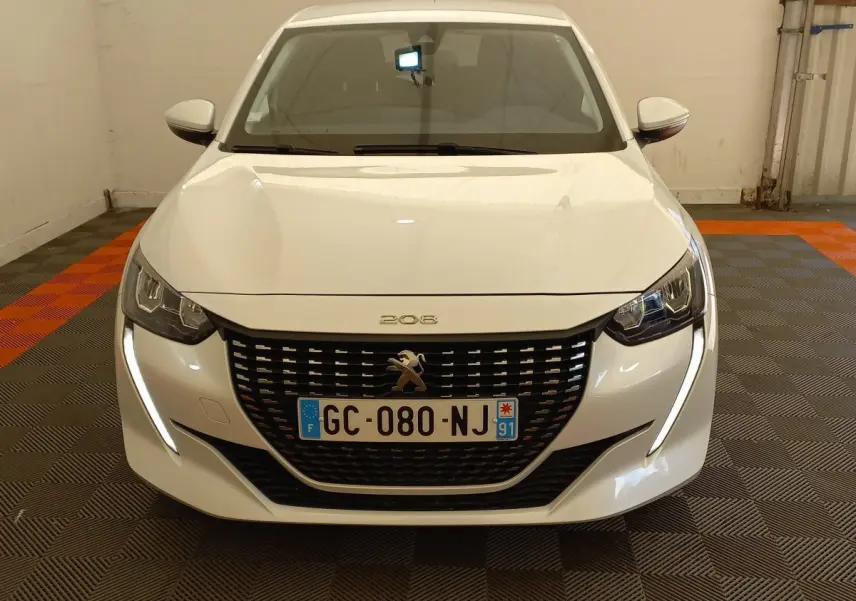 Vue frontale d'une Peugeot 208 blanche avec phares LED et calandre noire distinctive dans un garage.
